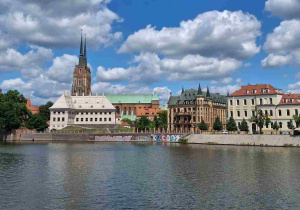 Szeroka panorama Ostrowa Tumskiego widziana z perspektywy rzeki Odry. Nad brzegiem wznoszą się zabytkowe kamienice i gmachy, nad którymi górują strzeliste wieże gotyckiej katedry.
