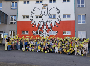 Uczniowie, nauczyciele podnoszą żółty balon do góry na tle Szkoły.