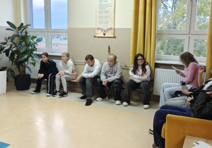 Uczennica czyta tekst o powstaniu książki z pergaminu. Uczniowie siedzą na sofie i na ławeczce i słuchają.