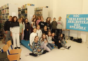 Uczniowie znajdują się w bibliotece i pozują do wspólnego zdjęcia. Nauczyciel biblioteki jest z uczniami.