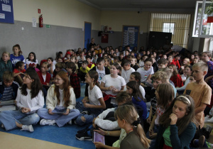 zdjęcie ukazuje grupę siedzących na korytarzu szkolnym dzieci wraz z nauczycielem, które rozwiązują quiz językowy