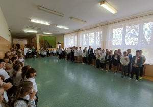 Korytarz szkolny. Uczniowie ustawieni klasami oczekują na rozpoczęcie uroczystości szkolnej z okazji Święta Niepodległości. W tle chłopiec trzyma w rękach flagę, obok niego dwie dziewczynki.