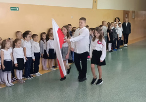 Korytarzem idzie uczeń trzymający w rękach flagę, obok niego dwie uczennice. W tle stoją uczniowie klas I - III.
