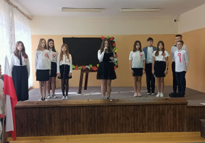 Scena szkolna. Na niej stoi stelaż z tablicą. Obok niej, z prawej i lewej strony stoją uczniowie. Jedna z uczennic trzyma w ręku mikrofon i czyta informacje dotyczącą Święta Niepodległości. Uczniowie mają przypięte kotyliony.