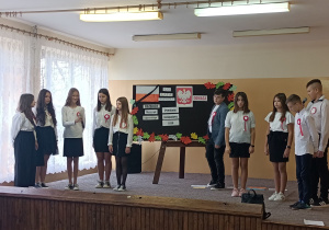 Scena, na niej uczniowie. Jeden z uczniów umieścił na tablicy napis „odwaga”, drugi przez mikrofon czyta tekst o odwadze Polaków.