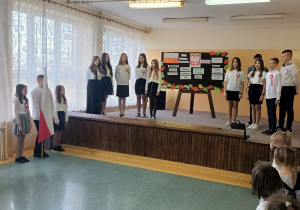 Scena, na niej występujący uczniowie klasy 6. Między nimi, w tle, tablica z hasłami: rozbiory Polski, powstanie listopadowe, powstanie styczniowe, Mazurek Dąbrowskiego, odzyskanie niepodległości.