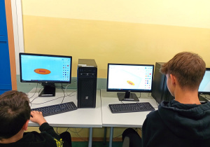 Pracownia komputerowa. Dwóch chłopców projektuje swoje imienne breloczki. Na pulpicie otwarte okno programu Tinkercad, w nim trójwymiarowe breloczki zaprojektowane przez chłopców.