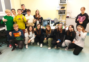 Pracownia komputerowa. Na biurku widoczna drukarkan3D. Obok biurka stoi pięciu chłopców i dwie dziewczynki. Przed biurkiem sześć uczennic siedzi na zgiętych kolanach. Wszyscy w dłoniach trzymają swoje zaprojektowane i wydrukowane imienne breloczki.