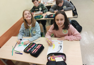 Sala lekcyjna, na ławkach szkolnych kredki i piórniki uczniów. Przy pierwszej ławce siedzą dwie dziewczynki, przy drugiej dwóch chłopców. Uczniowie kolorują rysunki według kolorów zakodowanych wynikami działań z tabliczki mnożenia.