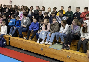 uczniowie siedzą na trybunach na hali sportowej i oglądają występ