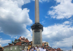 Uczniowie wraz z Panią Dyrektor stoją na schodach pod Kolumną Zygmunta III Wazy. Jest piękna pogoda. Uczniowie ubrani są na galowo.