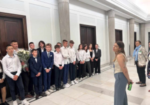 Uczniowie stoją na korytarzu łączącym Sejm z Senatem. Słuchają wykładu Pani przewodnik. Jeden z uczniów trzyma kwiaty, które zostaną wręczone w ramach podziękowania.