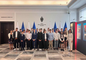 Uczniowie oraz nauczyciele ubrali w stroje galowe stoją w Senacie na tle ściany z napisem Senat Rzeczypospolitej Polskiej. Za uczniami znajdują się flagi Polski i Unii Europejskiej, a po lewej stronie drzwi do gabinetu Marszałka Senatu.