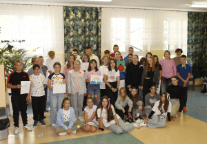 Duża grupa uczniów wraz z nauczycielkami znajdują się na korytarzu. Trzymają wykonane przez siebie rysunki oraz plakaty. Mają na sobie ubrania ze wzorami kropek. Niektórzy uczniowie mają we włosach kokardki w kropki .