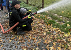 Chłopiec z grupy Żabki sprawdza swoje umiejętności gaśnicze przy użyciu węża strażackiego.
