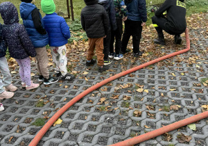 Przedszkolaki ustawione w rzędzie oraz strażacy przygotowujący stanowisko do obsługi węża gaśniczego.
