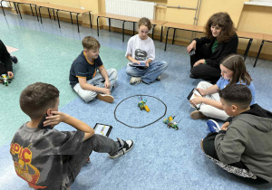 Uczniowie prezentują wyniki swojej pracy robotycznej przy pani bibliotekarce. Wszyscy znajdują się na korytarzu szkolnym.