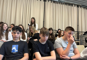 Uczniowie klas 8 siedzący na widowni, uczennica z mikrofonem zadaje pytanie radnym.