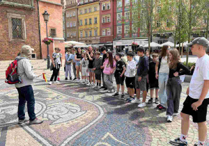 Grupa młodzieży stoi na zabytkowym, brukowanym rynku i słucha przewodnika opowiadającego o herbie Wrocławia. W tle kolorowe, wielopiętrowe kamienice z ogródkami gastronomicznymi.