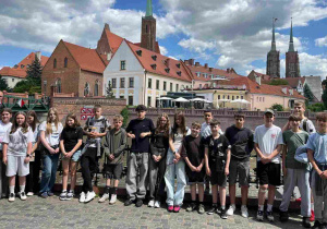 Grupa uczniów pozuje do wspólnego zdjęcia, stojąc na tle malowniczej panoramy Ostrowa Tumskiego. W tle widoczne są zabytkowe budynki z charakterystycznymi czerwonymi dachami oraz strzeliste wieże gotyckiej katedry.