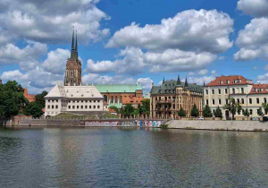 Szeroka panorama Ostrowa Tumskiego widziana z perspektywy rzeki Odry. Nad brzegiem wznoszą się zabytkowe kamienice i gmachy, nad którymi górują strzeliste wieże gotyckiej katedry.