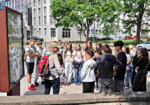 Uczniowie przed budynkiem Panoramy Racławickiej, skupiając się wokół przewodnika, który przekazuje im informacje.