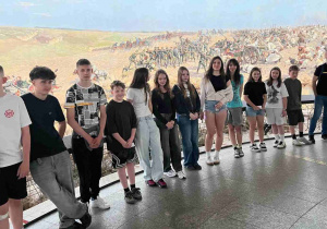 Uczniowie wraz z opiekunem na tarasie widokowym we wnętrzu Panoramy Racławickiej. W tle widać monumentalny obraz batalistyczny przedstawiający dynamiczną scenę bitwy z udziałem wojska i konnicy na otwartym polu.