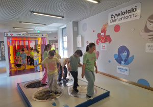 Uczniowie korzystają ze ścieżki sensorycznej , a za nimi na ściance Żywiołaki, które są maskotkami i motywem przewodnim strefy edukacyjnej.