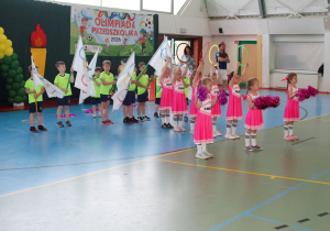 Układ choreograficzny w wykonaniu dzieci z grupy 5-6 latków: chłopcy machają flagami olimpijskimi natomiast dziewczynki w trzymają kółka olimpijskie i różowe pompony.