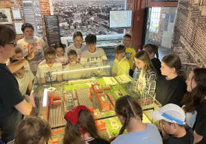 Uczniowie stoją wokół szklanej gabloty z makietą fabryki i słuchają przewodnika w muzeum o industrialnej historii Łodzi.