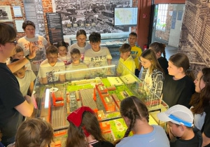 Uczniowie stoją wokół szklanej gabloty z makietą fabryki i słuchają przewodnika w muzeum o industrialnej historii Łodzi.
