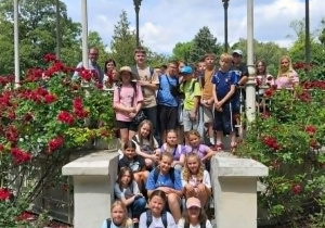 Grupa uczniów i opiekunów pozuje do zdjęcia na schodach zabytkowej, białej altany w parku otoczonym kwitnącymi, czerwonymi różami.