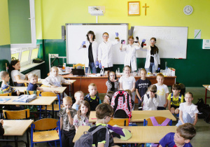 Uczniowie klasy 8a trzymają kolby zawierające wyniki doświadczenia a przed nimi młodsi uczniowie – obserwatorzy pokazów.