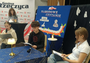 Uczennica klasy VII a czyta rolę Aniołka. Obok siedzą: uczeń, który za chwilę wcieli się w rolę Guślarza i uczennica, która wcieli się w rolę Chóru. Z tyłu przy pulpicie stoi bibliotekarka ze SP w Rzeczycy. Po jej prawej stronie na krzesełku siedzi i słucha Pani Dyrektor SP w Rzeczycy.