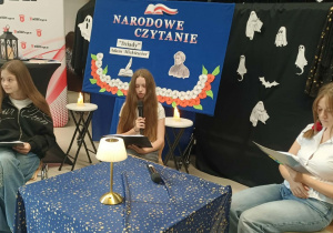 Uczennice SP w Rzeczycy siedzą przy stoliku. Jedna z nich czyta swoją rolę. Uczennica, która siedzi po jej prawej stronie delikatnie uśmiecha się do pozostałych uczestników Narodowego Czytania. Uczennica, która siedzi po jej lewej stronie patrzy w tekst.
