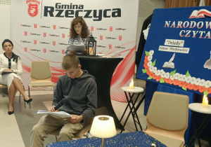 Bibliotekarka prowadząca biblioteki szkolne w Szkole Podstawowej w Rzeczycy czyta rolę narratora. Przy stoliku siedzi uczeń klasy VII a, który za chwilę będzie czytał rolę Guślarza. Po prawej stronie bibliotekarki siedzi i słucha Pani Dyrektor SP w Rzeczycy.