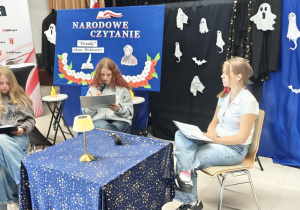 Uczennica klasy VIII a czyta rolę Dziewczyny 1. Obok niej, z prawej i lewej strony, siedzą jej koleżanki z klasy, które również biorą udział w Narodowym Czytaniu.
