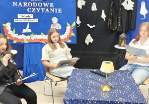 Uczennica klasy VII b czyta rolę Widma. Obok niej siedzą koleżanki z klasy VII a. W tle znajduje się dekoracja.