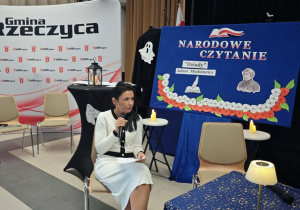Pani Dyrektor SP w Rzeczycy siedzi przy stoliku nakrytym ozdobnym obrusem. Za chwilę zacznie czytać swoją rolę. W tle znajduje się dekoracja.