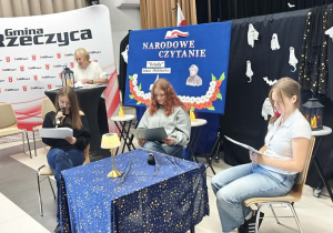 Uczennica kl. VIII a czyta rolę Dziewczyny 2. Obok niej siedzi koleżanka z klasy, która przed chwilą czytała rolę Dziewczyny 1. Przy stoliku siedzi również uczennica, która przed chwilą czytała rolę Chóru. Z tyłu stoi bibliotekarka z Biblioteki Publicznej w Rzeczycy. Po jej prawej stronie na krzesełku siedzi Pani Dyrektor SP w Rzeczycy.