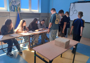 Uczniowie z klasy 6 stoją przy stoliku, uczennice z komisji wyborczej zapisują uczniów biorących udział w głosowaniu na Przewodniczącą Szkoły.