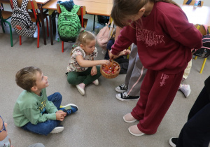 Uczennica z samorządu częstuje cukierkami z koszyka przedszkolaki siedzące na dywanie w sali.
