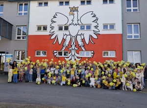 Uczniowie, nauczyciele podnoszą żółty balon do góry na tle Szkoły.