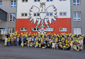 Uczniowie, nauczyciele podnoszą żółty balon do góry na tle Szkoły.