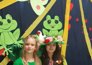 Przewodnicząca SU Maja ubrana na zielono i z zielonym kwiatem wpiętym we włosy,obok zastępca przewodniczącej SU Madzia ubrana w suknię w kwiaty, na głowie kapelusz. W tle wiosenna dekoracja: zielone żabki, motyl i duże uśmiechnięte słońce.