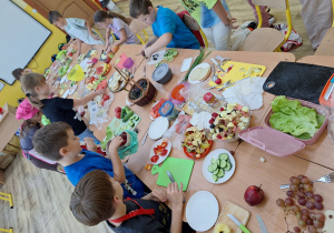 Stół zastawiony produktami przyniesionymi przez uczniów, uczniowie wybierają i kroją produkty.
