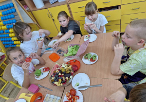 Uczniowie zajadają swoje udekorowane zdrowe kanapki a na środku stoi zrobiona sałatka owocowa.