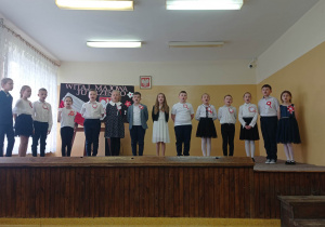 zdjęcie ukazuje grupę ubranych w białe bluzki dzieci, które stoją na scenie i śpiewają.