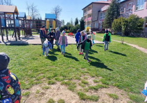 na zdjęciu widzimy grupę dzieci, ubranych w kurtki, jest słoneczny dzień. Dzieci sprzątają plac zabaw przed szkoła.
