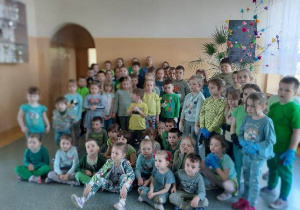 na zdjęciu widzimy grupę uśmiechniętych dzieci ubranych w zielone stroje. Dzieci stoją na korytarzu szkolnym.